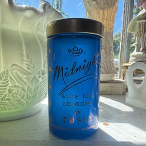 Op de afbeelding: Een cilindrische, kobaltblauwe eau de cologne fles met een zwarte dop. De fles heeft gouden tekst met de tekst "Midnight Blue Ice Cologne by Tussy". Gouden sterren en een kroonontwerp zijn ook aanwezig. De fles staat op een wit oppervlak.