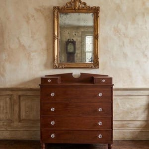 May include: A rectangular wall mirror with an ornate gold-toned frame featuring intricate carvings and a distressed glass surface, positioned above a dark brown wooden chest of drawers with six drawers and clear glass knobs on each drawer front.