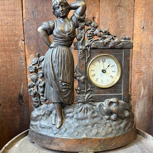 May include: Vintage bronze-effect clock featuring a woman figure, grape vines, and a clock face with Roman numerals. The clock has a detailed, aged appearance. The clock is set on a decorative base, suggesting it is a vintage piece.