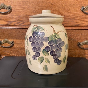 May include: A beige ceramic cookie jar with a lid, featuring hand-painted purple grapes and green leaves. The jar has a rounded shape and a matching lid. The design is a classic, country-style motif.