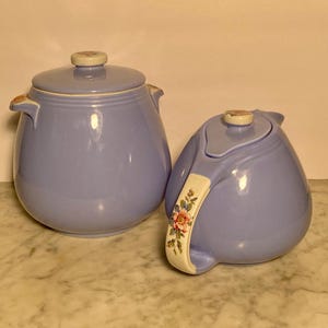 May include: A set of two vintage blue ceramic teapots with floral accents. The teapot on the left has a lid and two handles. The teapot on the right has a lid and a single handle with a floral design.