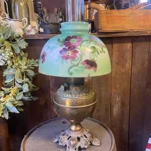 May include: A vintage oil lamp with a green glass shade featuring a floral design in shades of pink and purple. The lamp has a brass base with ornate details and sits on a wooden surface.
