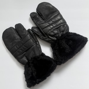 May include: A pair of black leather mittens with a split design for the index finger and thumb. The mittens have a quilted pattern and are trimmed with black faux fur around the wrists. The background is plain white.
