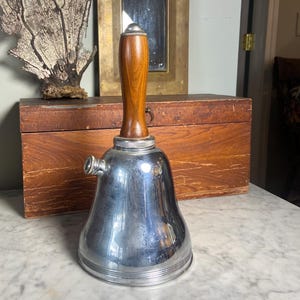 Può includere: Uno shaker per cocktail vintage in argento con un manico in legno. Lo shaker ha la forma di una campana e ha un beccuccio laterale.