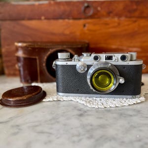 Op de afbeelding: Een vintage zwart-zilveren camera met een gele lens, op een wit kanten kleedje. Een bruine leren etui en lensdop zijn ook aanwezig. De camera heeft een klassiek ontwerp.