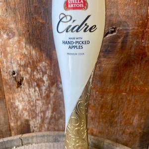 以下が含まれることがあります： 金色のアクセントが付いた白いステラアルトワシードルのタップハンドル。ハンドルにはステラアルトワのロゴと「Cidre」の文字が書かれています。「Made with Hand-Picked Apples」の文字も表示されています。