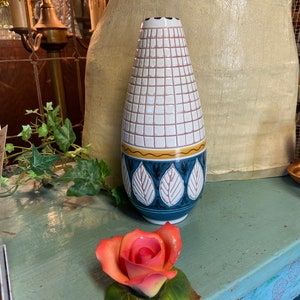 Puede incluir: Un jarrón de cerámica blanca con un patrón de cuadrícula y un diseño de hojas azules y amarillas. El jarrón está sobre una superficie azul con una rosa rosa.