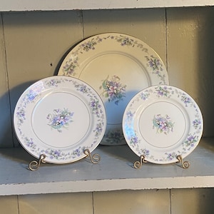 May include: Three white ceramic plates with a floral design featuring purple and white flowers. The plates are on gold metal stands.
