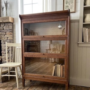 May include: A three-tiered, antique wooden bookcase with glass doors. The bookcase is a warm brown color and has a classic design. Books are visible inside the bottom shelf. The bookcase is in a room with a fireplace and a white chair.
