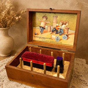 May include: A wooden sewing box with a hinged lid featuring a vintage illustration of children and spools of thread. The interior includes vertical wooden pegs, a rectangular burgundy velvet pincushion with O.N.T. text, and a small black pincushion.