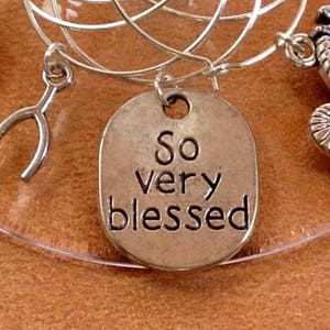 May include: A silver charm with the words "So very blessed" engraved on it. The charm is attached to a silver wire with other charms.