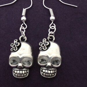 May include: A pair of silver-toned skull earrings with a small flower accent on each skull. The earrings have a detailed design, including visible teeth and eye sockets. The earrings hang from silver-colored ear wires.