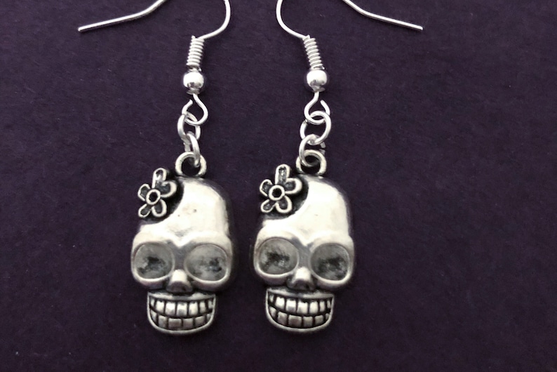 May include: A pair of silver-toned skull earrings with a small flower accent on each skull. The earrings have a detailed design, including visible teeth and eye sockets. The earrings hang from silver-colored ear wires.