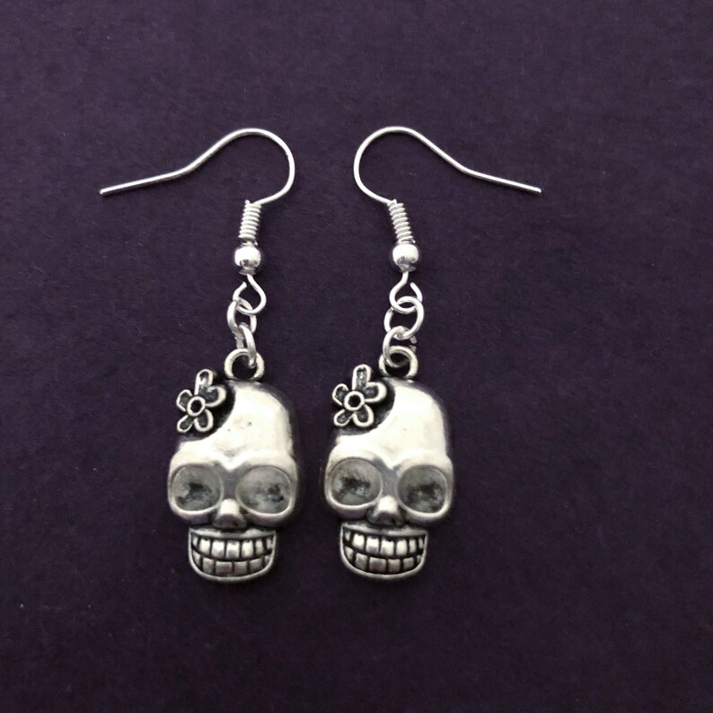 May include: A pair of silver-colored skull earrings with a flower accent on each skull. The earrings have a hook closure and are displayed against a dark purple background. The skulls have detailed features, including eye sockets and a grinning mouth.