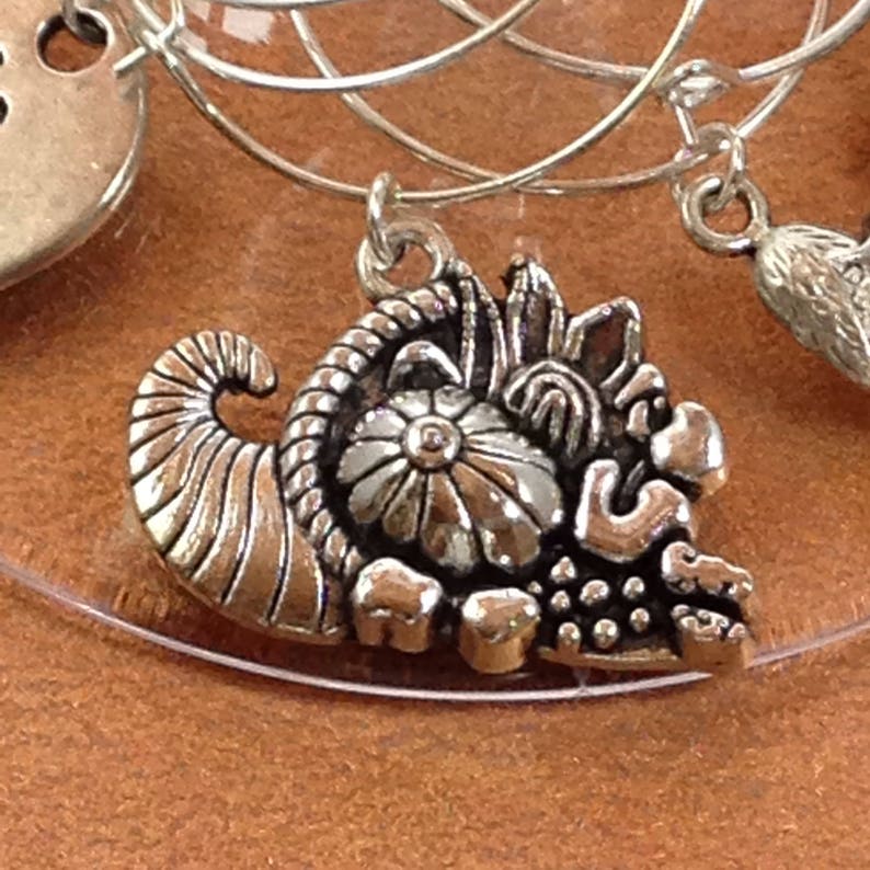 May include: Silver charm with a cornucopia design. The cornucopia is filled with grapes, a flower, and other decorative elements.