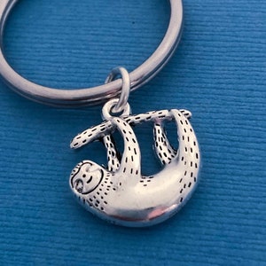 May include: A silver sloth keychain hanging from a silver key ring. The sloth is depicted hanging upside down from a branch.
