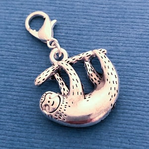 May include: Silver charm of a sloth hanging upside down from a branch. The charm has a lobster clasp for attaching to a bracelet or necklace.