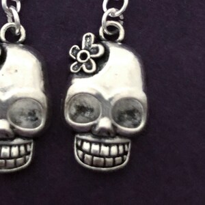 May include: Silver-toned skull earrings with a flower accent. The earrings feature a detailed skull design with a wide grin and large eye sockets. The skull on the right has a small flower on top. The earrings hang from a silver chain.
