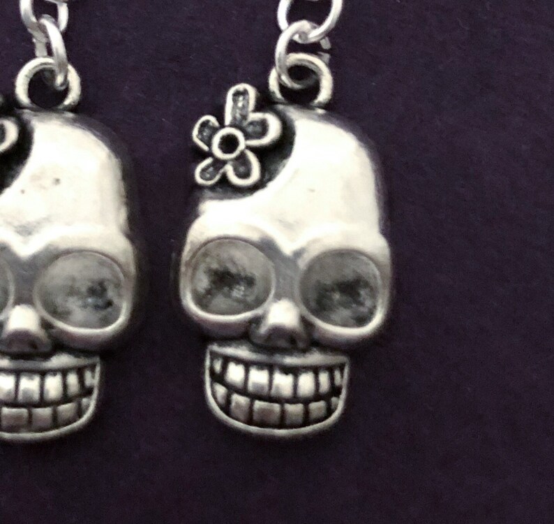 May include: Silver-toned skull earrings with a flower accent. The earrings feature a detailed skull design with a wide grin and large eye sockets. The skull on the right has a small flower on top. The earrings hang from a silver chain.