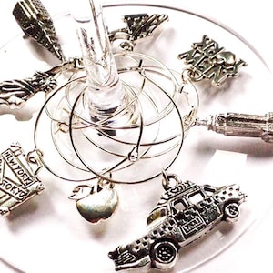 May include: Set of six silver-toned wine glass charms featuring New York City landmarks. The charms include the Statue of Liberty, the Empire State Building, a taxi cab, an apple, a sign that says "New York, Paris, Tokyo", and a sign that says "I Love NY".