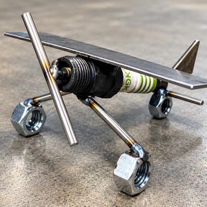 May include: A metal sculpture of an airplane constructed from a spark plug, steel nuts, and flat metal bars. The body is a spark plug with green and white branding, featuring a flat wing, a tail fin, and two steel nuts serving as wheels.