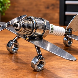 May include: A decorative airplane sculpture constructed from metal hardware, featuring an NGK spark plug as the main fuselage, metal nuts as wheels, and flat metal pieces forming the wings, propeller, and tail fin, with visible welded joints.
