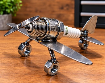 May include: A decorative airplane sculpture constructed from metal hardware, featuring an NGK spark plug as the main fuselage, metal nuts as wheels, and flat metal pieces forming the wings, propeller, and tail fin, with visible welded joints.