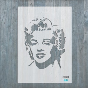 May include: A white stencil of Marilyn Monroe's face with a simple outline. The stencil is on a gray wood background. The text "CREATE Cuts" is in blue at the bottom of the stencil.