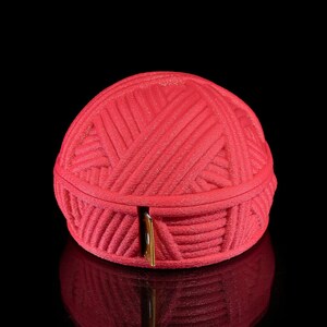 Pu&ograve; includere: Un contenitore rosso a forma di palla di filo con una chiusura color oro. Il contenitore &egrave; realizzato in un materiale strutturato e poggia su una superficie nera riflettente. Il contenitore &egrave; progettato per assomigliare a un gomitolo di filo.