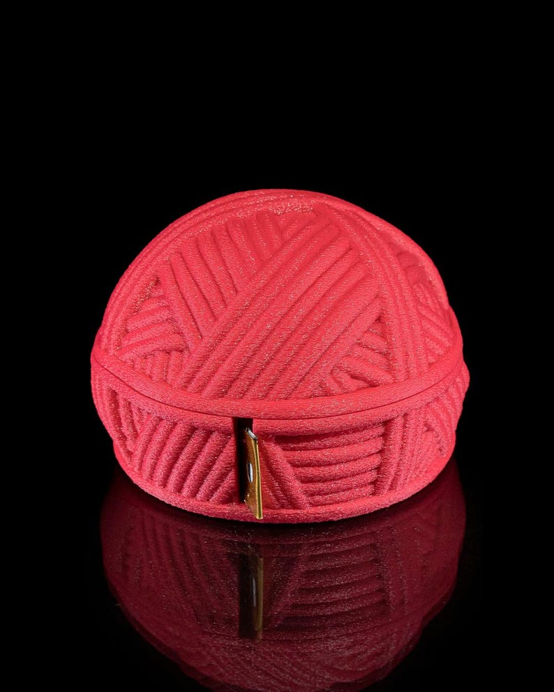 Pu&ograve; includere: Un contenitore rosso a forma di palla di filo con una chiusura color oro. Il contenitore &egrave; realizzato in un materiale strutturato e poggia su una superficie nera riflettente. Il contenitore &egrave; progettato per assomigliare a un gomitolo di filo.