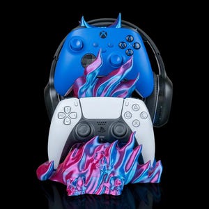 May include: A blue Xbox controller with horns sits above a white PlayStation 5 controller. Both are set on a flame-shaped stand with skulls, in shades of pink, blue, and purple. A black headset is behind the controllers.
