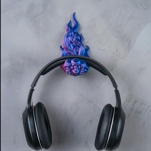 May include: A black pair of headphones hanging on a wall-mounted headphone holder. The holder is shaped like a flame with a skull design, featuring blue and pink hues. The headphones have a silver trim.