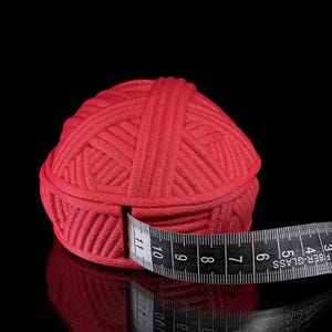 Pu&ograve; includere: Un gomitolo di lana rosso vivo viene misurato con un metro a nastro bianco e nero. Il nastro &egrave; avvolto attorno alla lana, mostrando le misure in centimetri. Il gomitolo poggia su una superficie nera riflettente.