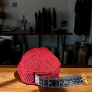Pu&ograve; includere: Un gomitolo di lana rosso vivo con un metro a nastro bianco e nero avvolto attorno. Il nastro mostra le misure in centimetri. Il gomitolo &egrave; su una superficie di legno, suggerendo un progetto artigianale o di cucito.