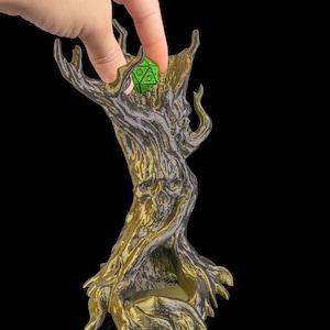 Puede incluir: Un detallado soporte para dados en forma de árbol con una calavera tallada en el tronco. El soporte está pintado en tonos dorados y grises, con un dado verde que se coloca dentro. El diseño es intrincado y de temática fantástica.