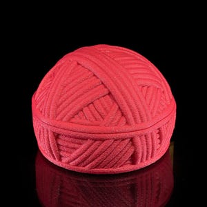 Pu&ograve; includere: Un contenitore rosso a forma di gomitolo con coperchio. Il contenitore &egrave; realizzato in un materiale strutturato, con l'aspetto di filato avvolto strettamente. Il contenitore &egrave; su una superficie nera riflettente.