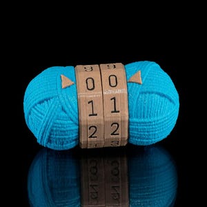 Puede incluir: Una bola de hilo turquesa vibrante con una cerradura de combinación de madera que muestra los números 00, 11 y 22. La bola de hilo se refleja en una superficie negra.
