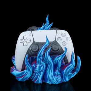May include: A white game controller rests on a blue flame-like base. The controller has black accents and button symbols. The base has a swirling design with hints of red. The background is black.