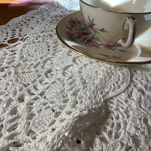 May include: A white crocheted doily with a floral pattern, a white teacup with a gold rim, and a matching saucer with pink floral designs.