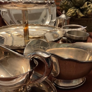 May include: A collection of antique silver serving pieces, including a tiered server, gravy boats, and serving spoons. The pieces have a reflective, polished finish, and are arranged on a brown surface. The collection suggests a formal dining setting.