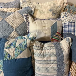 May include: A collection of various blue and white quilted pillows. The pillows feature different patterns, including floral, plaid, and geometric designs. The fabrics are a mix of solid and patterned cotton, creating a cozy, vintage aesthetic.