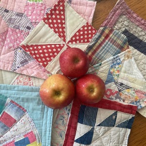 May include: A collection of colorful patchwork quilt squares with a red, white, and blue star design. Three red apples are placed on top of the squares.