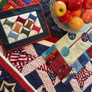 May include: A collection of handmade quilts and quilted items, featuring patriotic and geometric designs. The quilts showcase a variety of colors, including red, white, and blue, with star and flag motifs. A glass bowl of apples sits nearby.