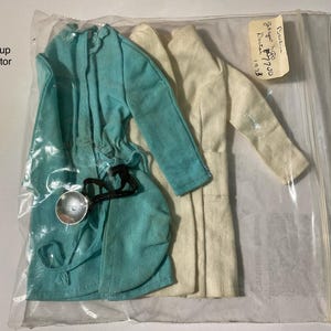 May include: A turquoise coat and a white coat for a doll, both with long sleeves, enclosed in a clear plastic bag. A small silver-colored stethoscope with a black cord is attached to the turquoise coat. The text Getup Doctor is visible.