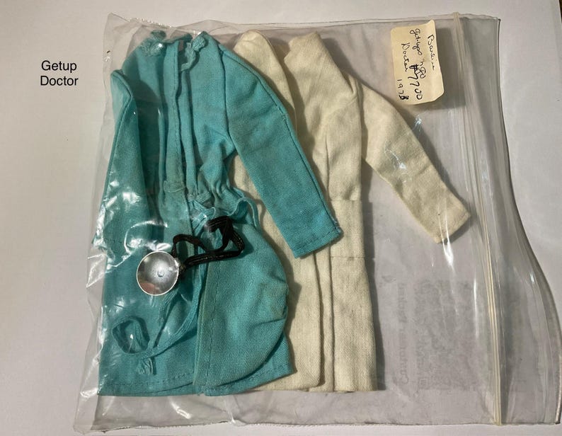 May include: A turquoise coat and a white coat for a doll, both with long sleeves, enclosed in a clear plastic bag. A small silver-colored stethoscope with a black cord is attached to the turquoise coat. The text Getup Doctor is visible.
