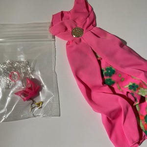 May include: A bright pink doll dress featuring a gold-tone circular accent at the neckline and a floral patterned underskirt. Beside the dress is a clear plastic bag containing a small pink doll shoe and a tiny gold-tone metal accessory.