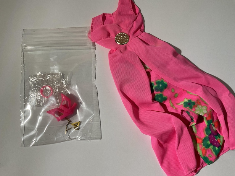 May include: A bright pink doll dress featuring a gold-tone circular accent at the neckline and a floral patterned underskirt. Beside the dress is a clear plastic bag containing a small pink doll shoe and a tiny gold-tone metal accessory.
