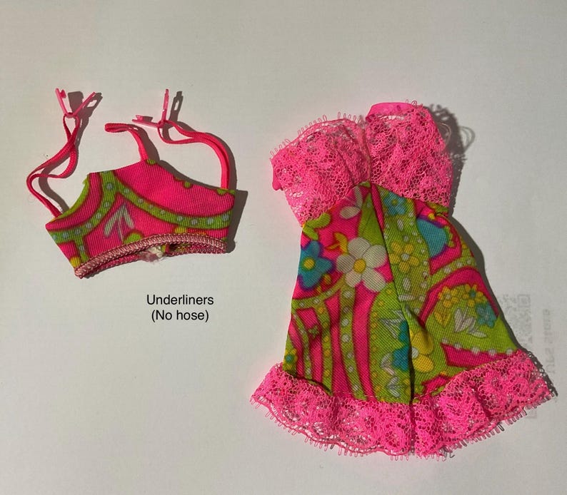 May include: A doll-sized outfit consisting of a pink and green floral patterned top with thin straps and a matching dress featuring a pink lace bodice and hem. The text "Underliners (No hose)" is printed below the top piece of the clothing set.