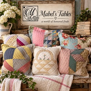 May include: A collection of six square quilted throw pillows featuring various patchwork patterns, floral prints, and a central basket motif. The pillows are arranged on a wooden surface with a sign reading Mabel's Tables a world of honored finds.