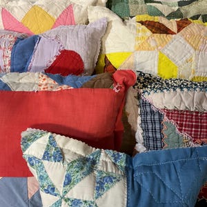 May include: A collection of rectangular throw pillows featuring various patchwork quilt designs. The pillows display diverse patterns including floral prints, solid red fabric, and geometric star motifs in colors such as yellow, blue, white, and green with visible quilted stitching throughout.